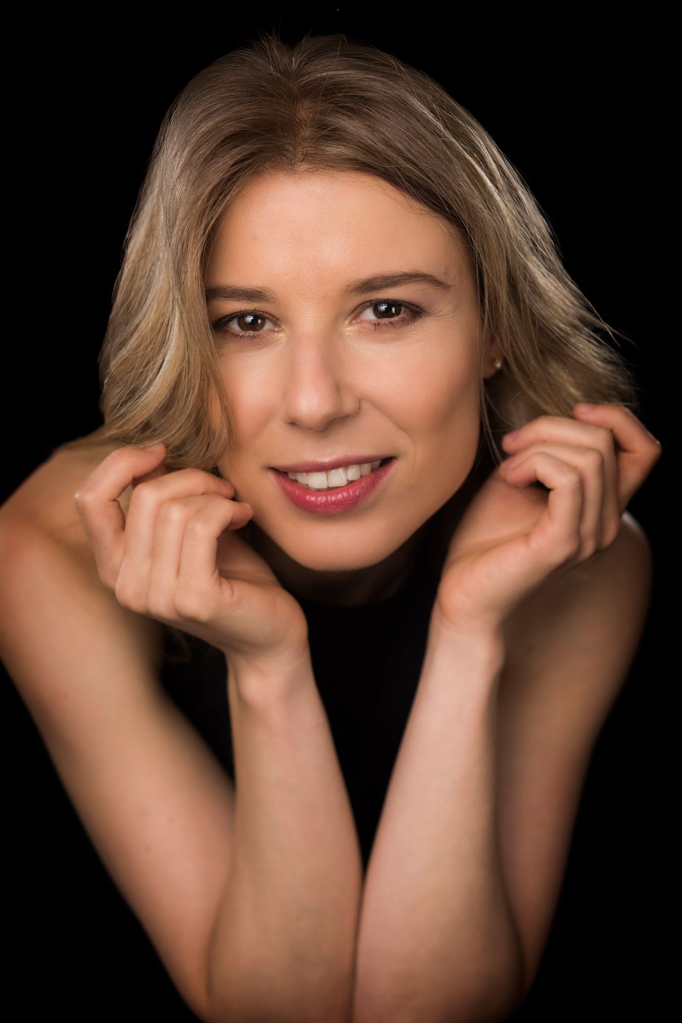 A woman smiling with both hands framing her face, in warm studio light
